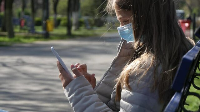 Sick Child with Protection Mask Playing on Smartphone, Sad Girl Using Smart Phone on Bench in Park, Kid in Coronavirus Pandemic