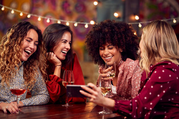 Multi-Cultural Group Of Female Friends Enjoying Night Out In Bar Looking At Mobile Phone