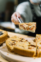 Unrecognisable woman taking spatula with piece of tasty open plum pie from shelf with different kinds of pies at cafe. Cafe, city lifestyle, food, dessert concept. Close up, selective focus, vertical.