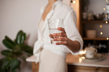 Unrecognizable woman in white shirt holding a glass of water, standing in the kitchen with evening light. Healthy lifestyle - drinking enough water every day 