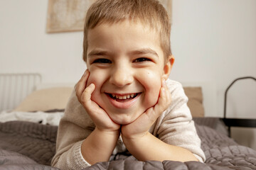 Portrait of little, smiling caucasian boy on the bed at home. Cute child relaxing, resting in bedroom. Positive emotions. Cozy and modern interior. Natural, earth colors. Casual clothes.