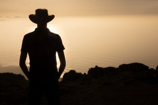 Silhouette of young man with hat against sunset, overlooking clouds and sea, warm colors, torso, copy space on the right