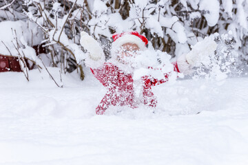 Happy small girl in red Christmas elf robe and hat with white gloves sitting in the snow and throwing it. Christmas card with little Santa’s helper in snow. Santa Claus little Scout Elf in the snow