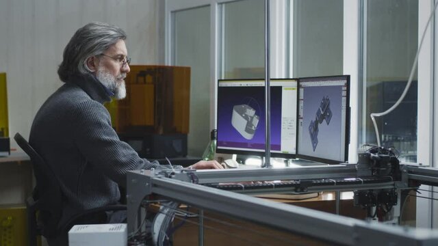 Mature man with gray hair and bread sitting at desk and creating 3D model on computer near coworker in modern design studio