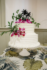 White classical two-tiered wedding cake with fresh pink flowers and greenery decoration on the white stand on the table Background of green and gold monstera leaves tablecloth 