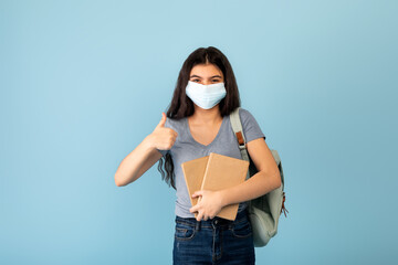 Funky Indian teen student in face mask with backpack holding books and showing thumb up gesture on blue background