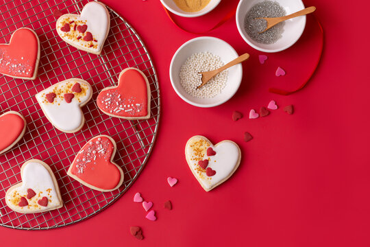 Heart-Shaped Cookies on a Cooling Rack Decorated with Red and White Icing; Bowls of Sprinkles; Red Background