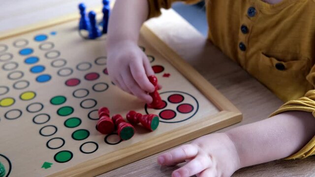 close-up of hands of small child manipulate with wooden figurines of family board game, smart kid, 2-year-old girl is learning to count, Cross and circle game Parcheesi, foreground focus