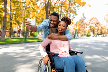 Cheerful black guy hugging his disabled girlfriend in wheelchair, making selfie on smartphone at autumn park
