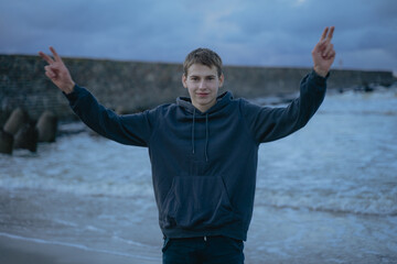 a guy in a hoodie stands at the sea shows two fingers