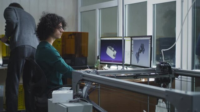 Young man creating 3D model on computer while sitting at desk near colleague using printer in modern design studio