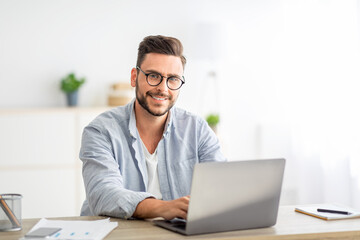 Portrait of freelancer man sitting at desk with laptop computer at home office, smiling at camera,...