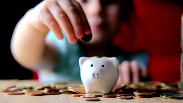close-up of hands of small child manipulate with metal coins, kid counts, puts euro union money in white piggy bank, concept of pocket money, life insurance, save up for gift, foreground focus