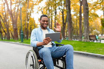 Impaired black man in wheelchair using tablet computer for remote communication at autumn park,...