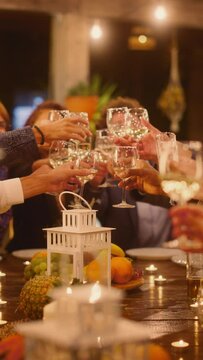 Vertical Screen: Big Dinner Party with a Small Crowd of Friends Celebrating at a Restaurant. Beautiful Happy Hosts Propose a Toast and Raise Wine Glasses while Sitting at a Table in the Evening.