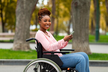 Happy disabled black woman in wheelchair using cellphone, checking social media, browsing web at autumn park