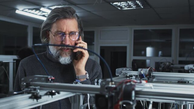 Mature bearded man in turtleneck and glasses talking with client on cellphone and watching modern printer printing 3D model in professional design studio
