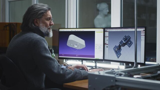 Middle aged man in sweater and glasses creating 3D models of details on computers while sitting at table and working in design workshop