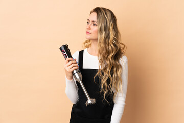 Young brazilian woman using hand blender isolated on beige background looking to the side