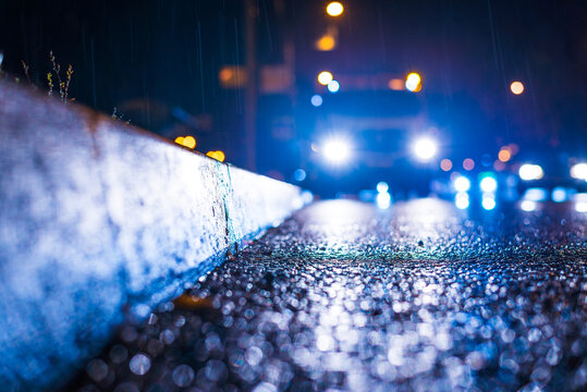 Night city in the rain.  Headlights of approaching cars. Reflections of light on a wet road. Falling drops of rain. Colorful colors. Focus on the asphalt. Close up view from the curb level.