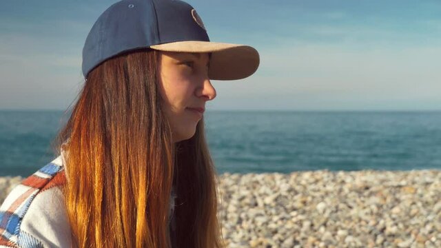 A woman in a baseball cap holds a teacup and thoughtfully looks deep into herself on a sunny day by the sea. The concept of lifestyle, self-acceptance, distribution of thoughts in the head.