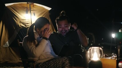 Excited Asian couple watching sport competition on the smartphone while camping at night
