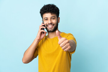 Young Moroccan man isolated on blue background keeping a conversation with the mobile while doing...