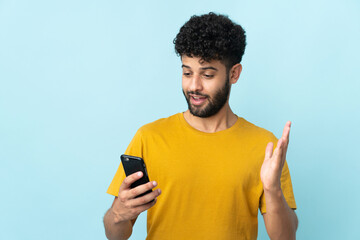 Young Moroccan man isolated on blue background looking at the camera while using the mobile with surprised expression