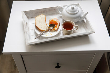 A teapot, a cup of tea, two slices of toasted bread with apricot jam on a tray on the bedside table.