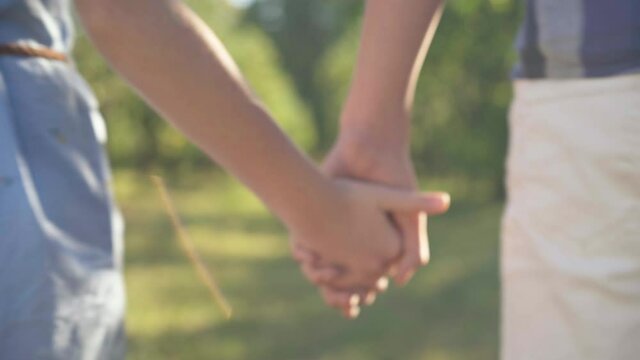 Close-up teenage couple taking hands and walking away in sunshine. Caucasian teen boy and girl holding hands strolling in summer spring park outdoors in sunshine. First love and adolescence concept