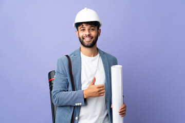 Young architect Moroccan man with helmet and holding blueprints over isolated background giving a...