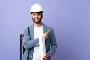 Young architect Moroccan man with helmet and holding blueprints over isolated background pointing...