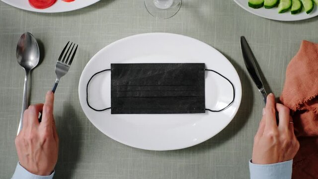 black medical mask on a white plate in a restaurant. a man sits at a table and holds a fork and a knife in his hands. the concept of coronovirus and restaurant business