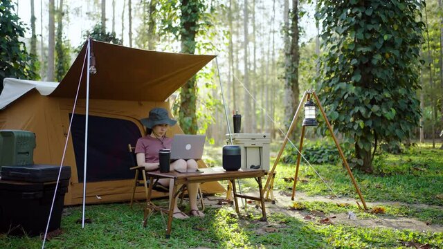 Asian woman using laptop working remotely while camping in the forest. work and vacation concept.