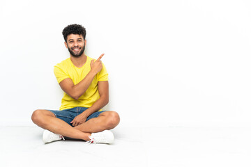 Fototapeta premium Young Moroccan handsome man sitting on the floor over isolated background pointing finger to the side