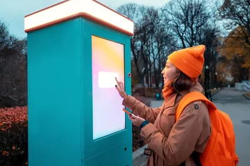 Deurstickers A woman uses a self-service kiosk to print photos from her smartphone on a city street © EdNurg