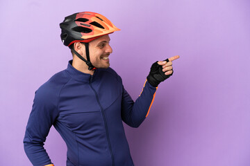 Young Brazilian cyclist man isolated on purple background pointing finger to the side and presenting a product - obrazy, fototapety, plakaty