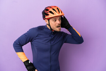 Young Brazilian cyclist man isolated on purple background listening to something by putting hand on the ear
