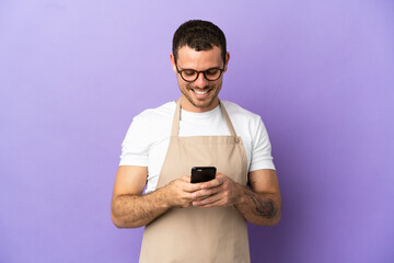 Brazilian restaurant waiter over isolated purple background sending a message with the mobile