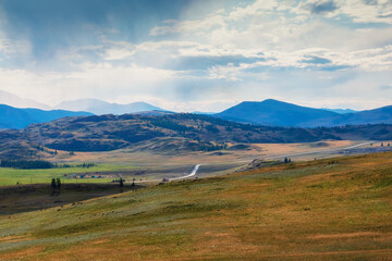 Rainy alpine view from green hills to high snow mountain range in sunlight during dramatic in changeable weather. Winding asphalt road among the hills.