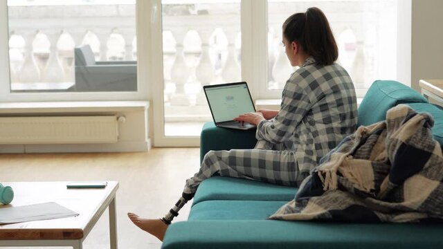 Back view of woman with artificial bionic high technology leg prosthesis working on laptop at home