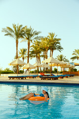 A woman in a sun hat in the pool in a swimming circle with a laptop works remotely against the background of palm trees and sun loungers.