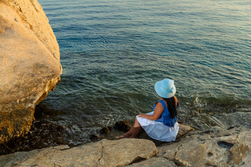 Fototapeta na wymiar A young woman in a white skirt and a sun hat on the seashore sits on a stone admiring nature with her back to the camera.