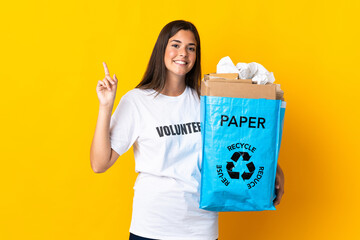 Young brazilian girl holding a recycling bag full of paper to recycle isolated on yellow background showing and lifting a finger in sign of the best