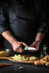 The chef cuts raw potatoes into pieces with a knife before preparing breakfast or dinner. Close-up of a cook hands while working in a restaurant kitchen.