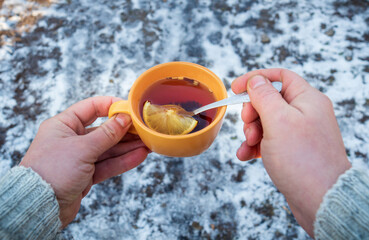Men hand hold an orange mug of hot herbal tea in cold winter. Prevention of viral diseases lemon tea. Spoon in person is hand.