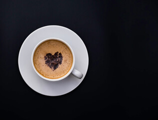 A mug of coffee with a chocolate heart on the foam stands on a monochromatic black background