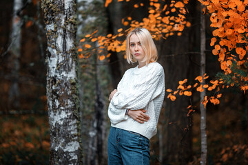 a tall young blonde in an autumn forest dressed in a white sweater and jeans