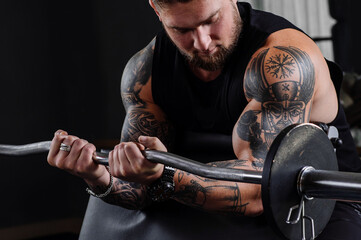 Tattooed bodybuilder does exercises and swings his biceps with a barbell in the gym
