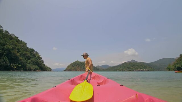 Man with sunglasses and hat rows pink plastic canoe along sea against green hilly islands with wild jungles. Traveling to tropical countries.